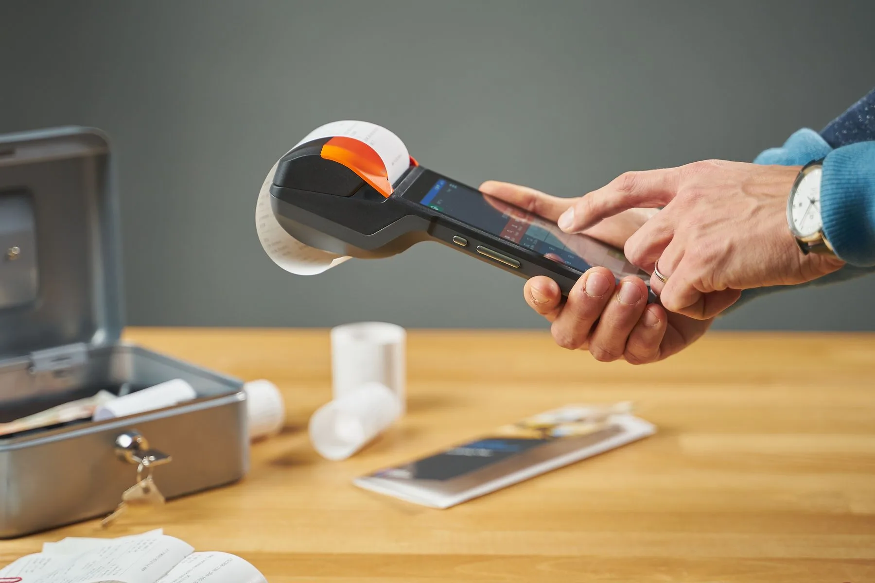 Haende bedienen einen mobilen Bondrucker, daneben liegen Kassenbelege, Bonrollen und eine Geldkassette